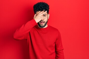 Wall Mural - Young arab man with beard wearing casual red sweater peeking in shock covering face and eyes with hand, looking through fingers with embarrassed expression.