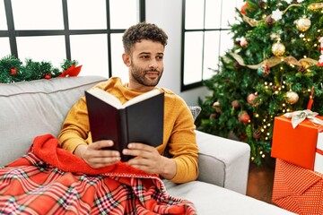 Canvas Print - Young arab man reading book lying on the sofa by christmas tree at home.