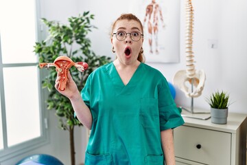 Poster - Young redhead doctor woman holding anatomical model of female genital organ scared and amazed with open mouth for surprise, disbelief face