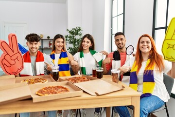 Wall Mural - Group of young hispanic people eating pizza supporting soccer team at home smiling happy pointing with hand and finger