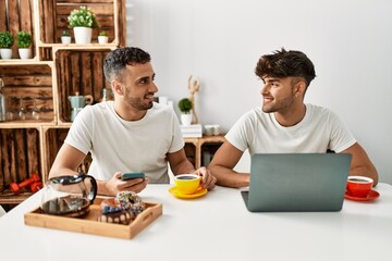 Wall Mural - Two hispanic men couple having breakfast using smartphone and laptop at home
