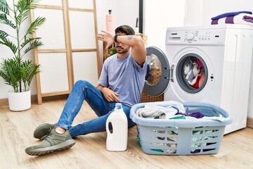 Wall Mural - Young hispanic man putting dirty laundry into washing machine covering eyes with arm, looking serious and sad. sightless, hiding and rejection concept