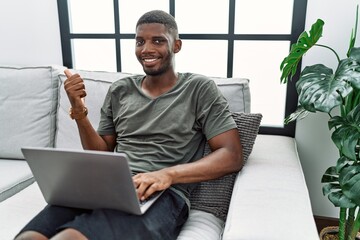 Canvas Print - Young african american man using laptop at home sitting on the sofa pointing to the back behind with hand and thumbs up, smiling confident