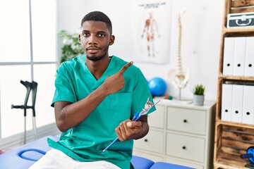 Poster - Young african american man working at pain recovery clinic pointing with hand finger to the side showing advertisement, serious and calm face