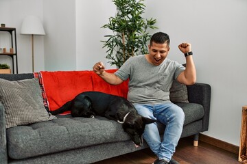 Wall Mural - Young latin man and dog sitting on the sofa at home dancing happy and cheerful, smiling moving casual and confident listening to music