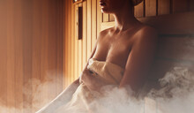 Young woman relaxing and sweating in hot sauna wrapped in towel. Girl In Sauna. Interior of Finnish sauna, classic wooden sauna with hot steam. Russian bathroom. Relax in hot sauna with steam