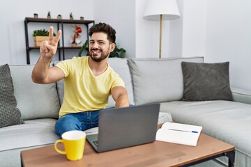 Sticker - Young man with beard using laptop at home smiling looking to the camera showing fingers doing victory sign. number two.