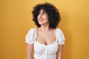 Poster - Young brunette woman with curly hair standing over yellow background smiling looking to the side and staring away thinking.