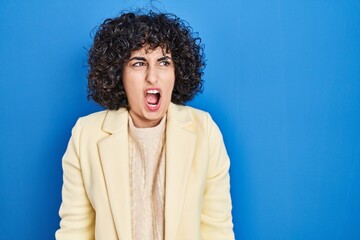 Poster - Young brunette woman with curly hair standing over blue background angry and mad screaming frustrated and furious, shouting with anger. rage and aggressive concept.