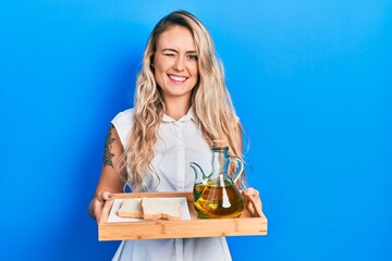 Poster - Beautiful young blonde woman holding tray with olive oil and bread toast winking looking at the camera with sexy expression, cheerful and happy face.