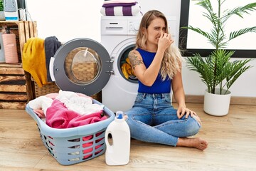 Poster - Young beautiful woman doing laundry sitting by wicker basket smelling something stinky and disgusting, intolerable smell, holding breath with fingers on nose. bad smell