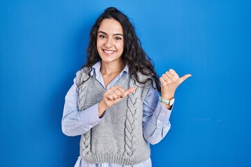 Sticker - Young brunette woman standing over blue background pointing to the back behind with hand and thumbs up, smiling confident