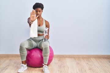 Wall Mural - African american woman wearing sportswear sitting on pilates ball doing stop sing with palm of the hand. warning expression with negative and serious gesture on the face.