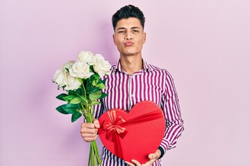 Wall Mural - Young hispanic man holding anniversary present and bouquet of flowers looking at the camera blowing a kiss being lovely and sexy. love expression.