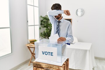 Poster - Hispanic man with beard voting putting envelop in ballot box smiling cheerful playing peek a boo with hands showing face. surprised and exited