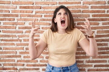 Sticker - Young brunette woman standing over bricks wall crazy and mad shouting and yelling with aggressive expression and arms raised. frustration concept.