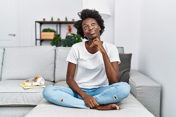 Sticker - African young woman sitting on the sofa at home with hand on chin thinking about question, pensive expression. smiling with thoughtful face. doubt concept.