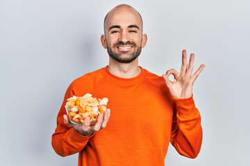 Wall Mural - Young bald man holding potato chip doing ok sign with fingers, smiling friendly gesturing excellent symbol