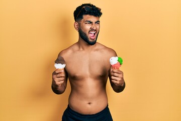 Wall Mural - Arab man with beard wearing swimwear eating two ice cream cones angry and mad screaming frustrated and furious, shouting with anger. rage and aggressive concept.