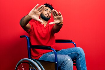 Poster - Arab man with beard sitting on wheelchair doing frame using hands palms and fingers, camera perspective