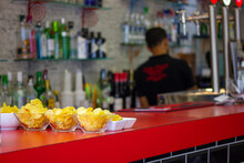 Piles, Plates, Bowls Of Potato Chips Tapas, A Traditional Spanish Snack That Is Served In Bars, Cafes, Restaurantes. Red Bar In A Tavern. Bartender In Defocus. Delicious Appetizer Starter Food, Meal.