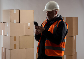 Poster - Warehouse manager. Man with phone in protective helmet. Warehouse manager in front of boxes. Warehouse manager uses mobile apps. Application for logistic accounting of concert. Guy in orange vest