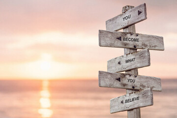 Wall Mural - you become what you believe text engraved on wooden signpost by the ocean during sunset.