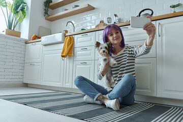 Wall Mural - Young woman carrying little dog and making selfie while sitting on the floor at the kitchen