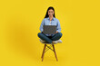 © Prostock-studio - Cheerful young chinese lady student sitting on chair and working on laptop, isolated on yellow background