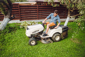 Wall Mural - Gardener driving a riding lawn mower in a garden. Professional landscaper using tractor at mowing lawn.