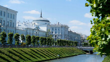 Beautiful Cityscape With A Narrow Canal And Two Slopes Covered By Green Grass On Blue Cloudy Sky Background. Stock Footage. Summer City Street With White Houses, And People Walking On The Bridge.