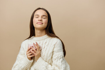 Wall Mural - a beautiful woman with loose hair is standing on a brown background in a knitted sweater, gently holding a mug in her hands, looking mysteriously to the side and smiling easily