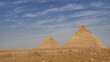 Two great pyramids of Cheops and Chephren on a background of blue sky and clouds. In the foreground is the sandy-rocky soil of the Giza plateau, a road curb. Egypt