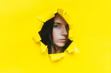 Close-up Portrait Of A Caucasian Young Woman Looking Through A Hole In Yellow Paper. An Incredulous Gaze. Women's Curiosity And Gossip. Jealous Wife. Spy Concept.