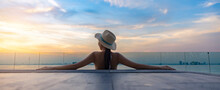 Asian Beautiful Woman In Hat Watching The Beautiful Scenic Sunset At The Edge Of The Infinity Pool With Cityscape, Enjoying Nature Background Banner Panorama. Woman From Behind Relaxing At Landscape.