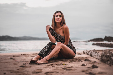 Wall Mural - Portrait of a brunette woman wearing a swimsuit and a pareo on the beach in summer, sitting on the sand