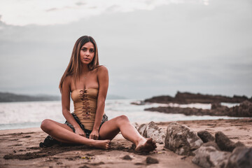 Wall Mural - Portrait of a brunette woman wearing a swimsuit and a pareo on the beach in summer, sitting on the sand