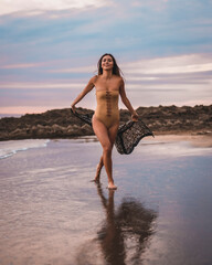 Wall Mural - A brunette woman having fun with a swimsuit and a pareo walking along the beach in summer at sunset