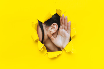 Wall Mural - Close-up of a left woman's ear and hand through a torn hole in the paper. Bright yellow background, copy space. The concept of eavesdropping, espionage, gossip and tabloids.