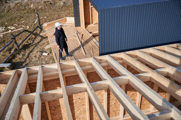 Wall Mural - Male engineer building wooden frame house. Man developer on construction site, inspecting quality of work on sunny day, holding hammer in hands.