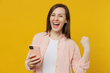 Wall Mural - Young happy excited woman she 30s wears striped shirt white t-shirt hold in hand use mobile cell phone chatting onlie browsing internet do winner gesture isolated on plain yellow background studio