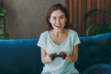 Young Happy Smiling Woman She 20s Wear Casual Clothes Mint T-shirt Hold In Hand Play Pc Game With Joystick Console Sit On Blue Sofa Indoor Rest At Home In Own Room Apartment. People Lifestyle Concept.
