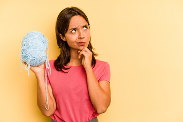 Wall Mural - Young hispanic woman holding a ball of wool isolated on yellow background looking sideways with doubtful and skeptical expression.