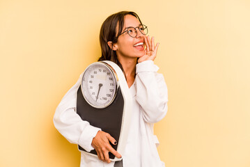 Wall Mural - Young nutritionist woman holding a weighing machine isolated on yellow background shouting and holding palm near opened mouth.