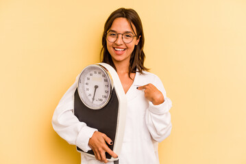 Wall Mural - Young nutritionist woman holding a weighing machine isolated on yellow background person pointing by hand to a shirt copy space, proud and confident