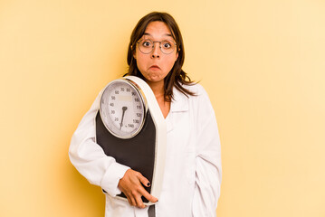 Wall Mural - Young nutritionist woman holding a weighing machine isolated on yellow background shrugs shoulders and open eyes confused.