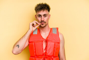 Wall Mural - Young caucasian man wearing a life jacket isolated on yellow background with fingers on lips keeping a secret.