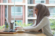 © wattana - Side view young businesswoman wearing hijab working with laptop in modern workplace.