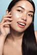 © Liubov Levytska - Vertical of beautiful asian woman with glowing perfect skin, applies daily skincare cream, cosmetic product on her cheek for clear, glowing face, blue background