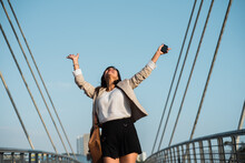 Working Woman Raises Her Hands In Happiness And Triumph On The Street, Holding A Cell Phone In Her Hand And Is Outdoors. Copy Space